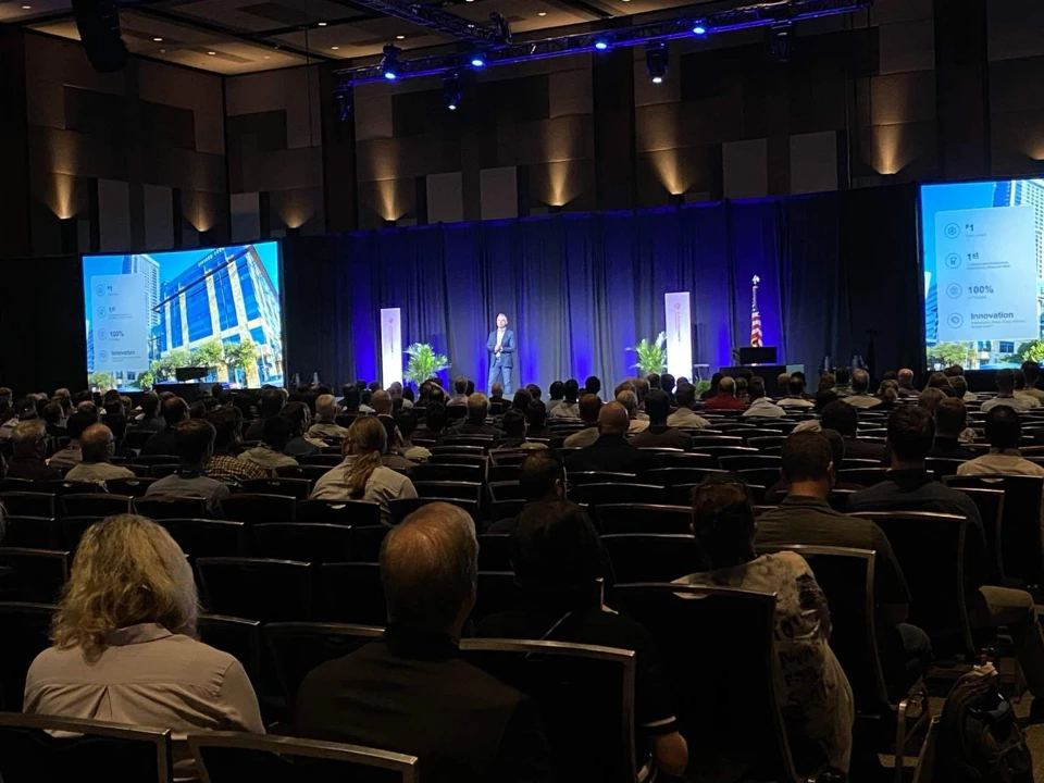 Conference hall with a speaker on stage, blue lighting, and two large screens. Audience seated, facing the stage, with an American flag in the background.
