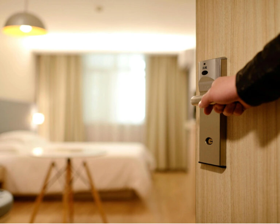 A person opening the door to a hotel room with a neatly made bed and soft lighting inside.