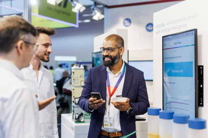 Three men talking at a trade fair booth displaying an environmental sensor with artificial intelligence.