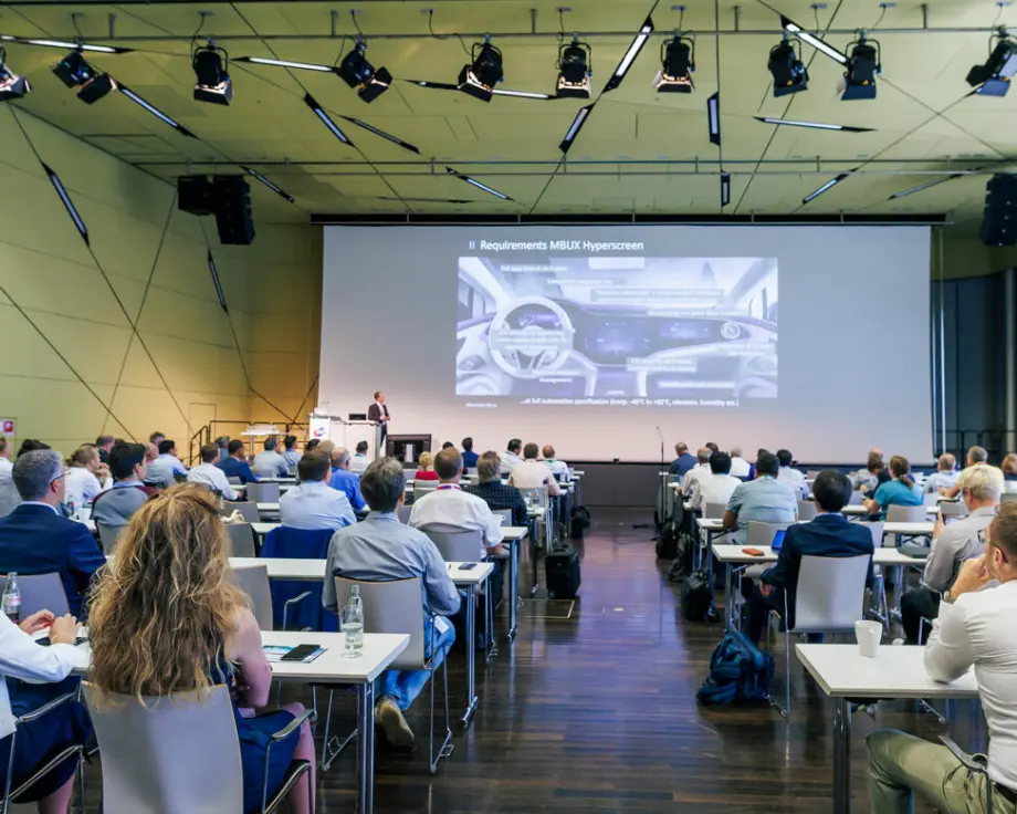 A conference room with attendees seated at tables, facing a speaker presenting about the "Requirements MBUX Hyperscreen" on a large screen. The audience is engaged, with some using laptops.